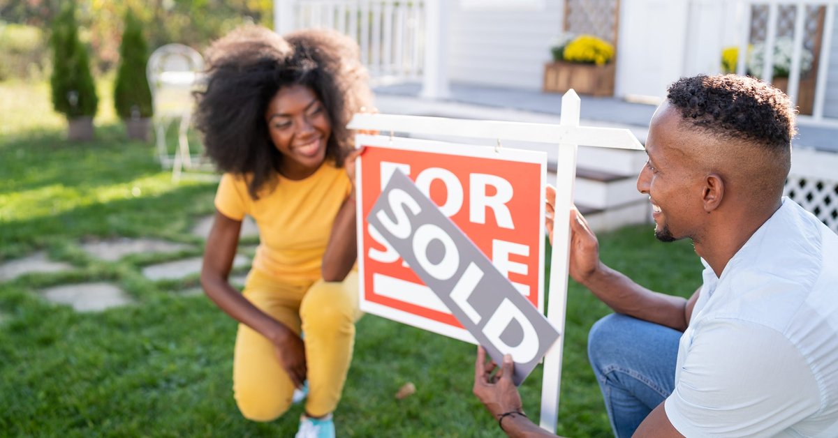 A young couple happily attaches a "Sold" banner to a "For Sale" sign outside a house.