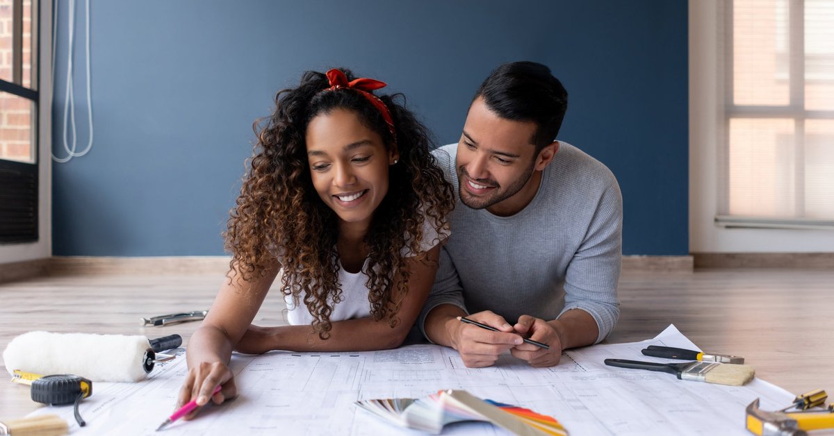 A couple looks at the blueprints of their new house while lying on the floor smiling.