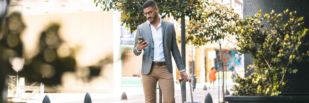 Man walking down street with luggage while looking at his cellphone.