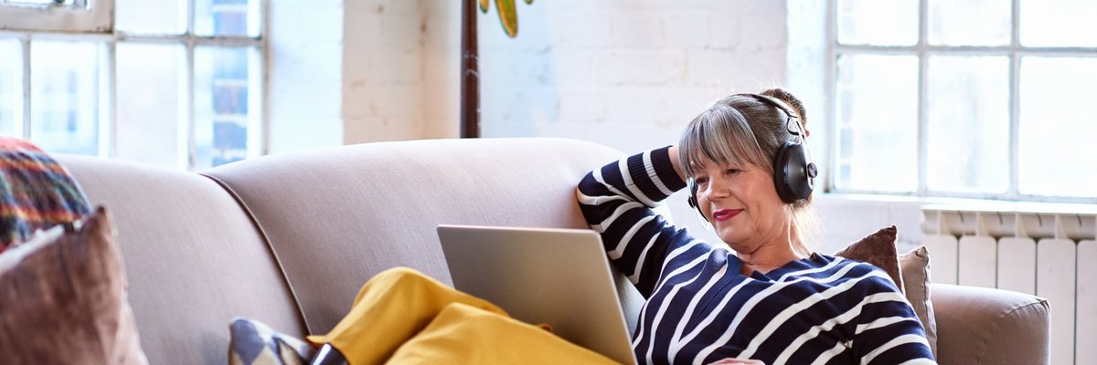 A person wearing headphones looks a their computer while lounging on the couch.