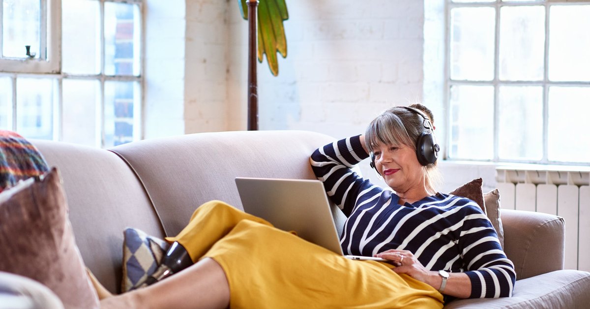 A person wearing headphones looks a their computer while lounging on the couch.