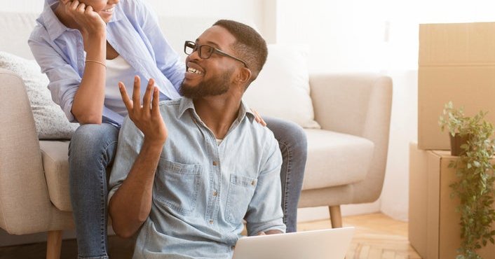 A couple sits in their living room, smiling as they look at a laptop.