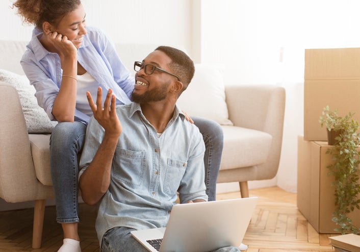 A couple sits in their living room, smiling as they look at a laptop.