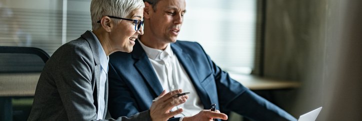 Two mature colleagues discuss while looking at a laptop in an office.