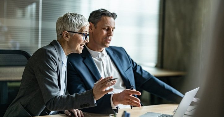 Two mature colleagues discuss while looking at a laptop in an office.