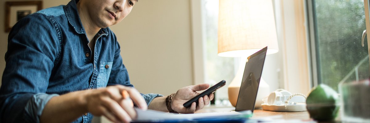 A person flips through papers at a home desk with a phone and laptop.