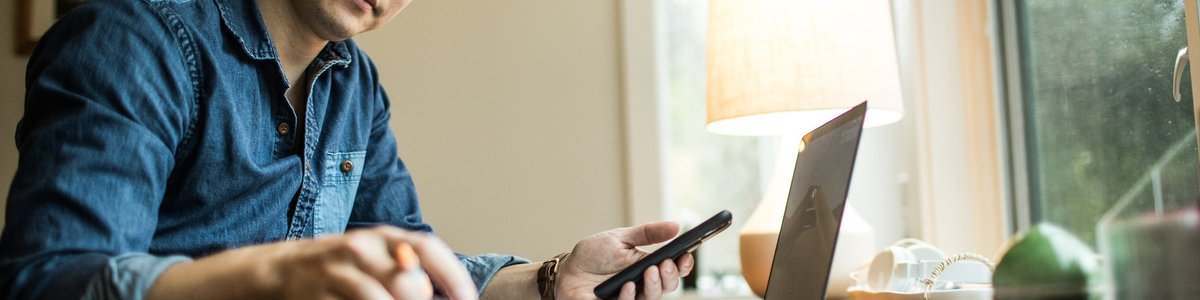 A person flips through papers at a home desk with a phone and laptop.