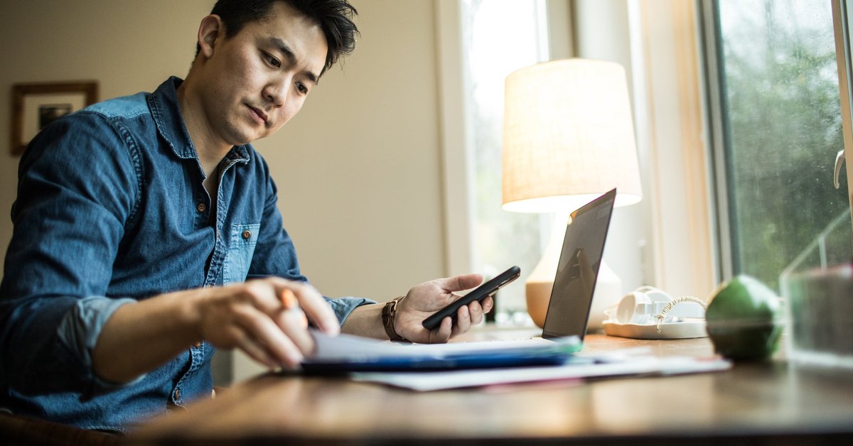 A person flips through papers at a home desk with a phone and laptop.