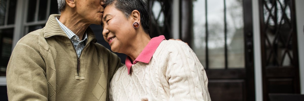 An older person kisses their partner's forehead as they embrace in front of a house.