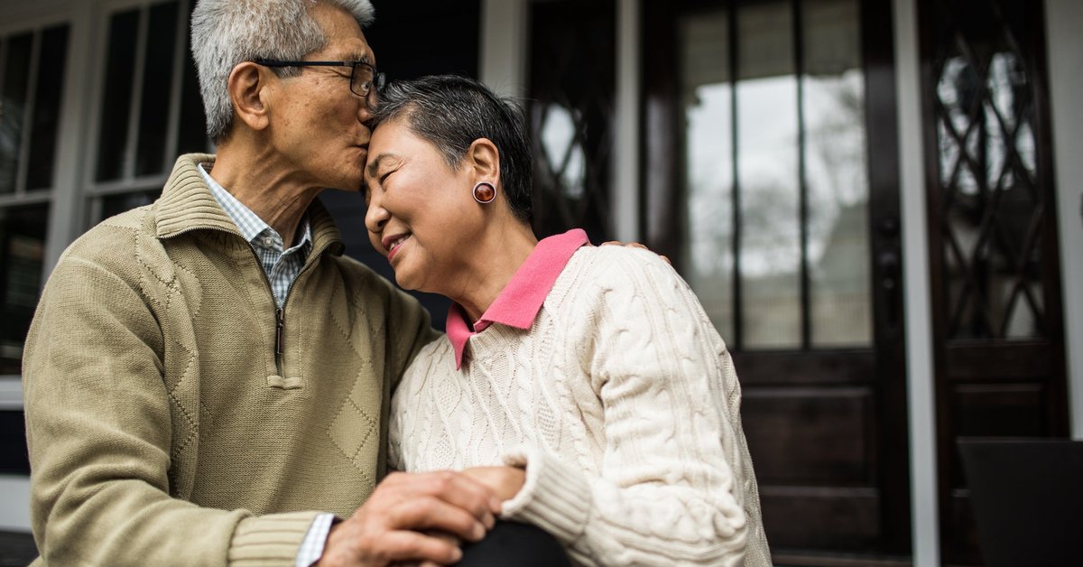 An older person kisses their partner's forehead as they embrace in front of a house.