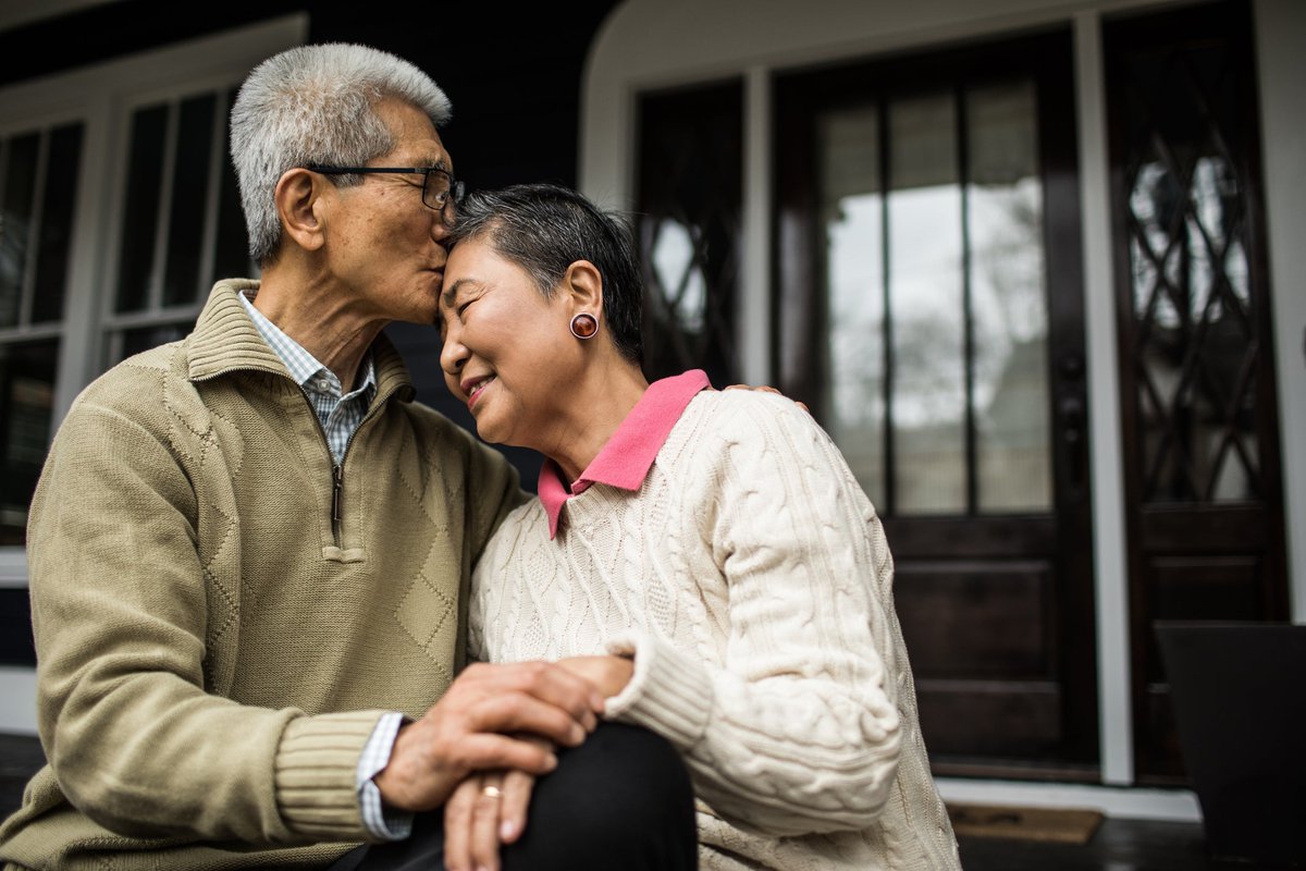 An older person kisses their partner's forehead as they embrace in front of a house.