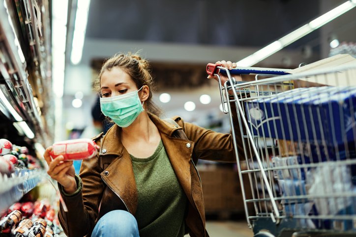 Woman wearing a mask examines groceries with one hand on her cart.