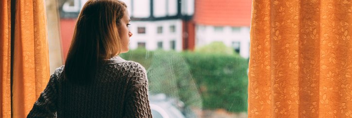 A woman looks out her window worriedly, holding a mug.