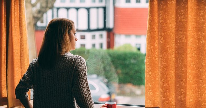 A woman looks out her window worriedly, holding a mug.