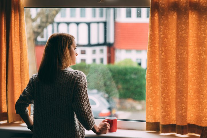 A woman looks out her window worriedly, holding a mug.