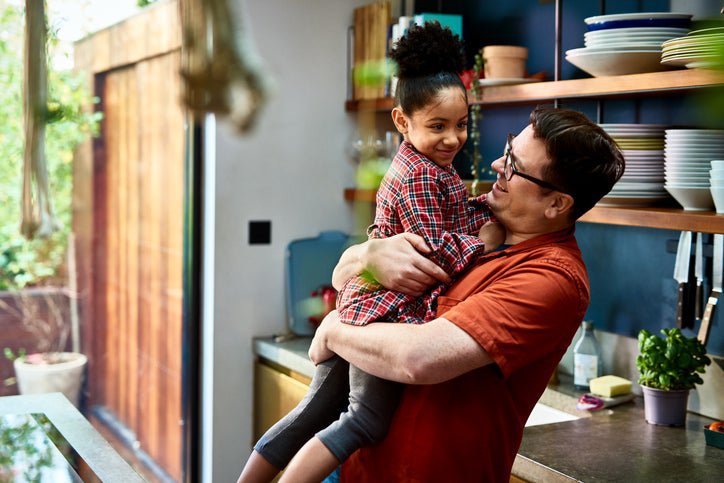 Parent holds their young child up in their kitchen. Both smile.