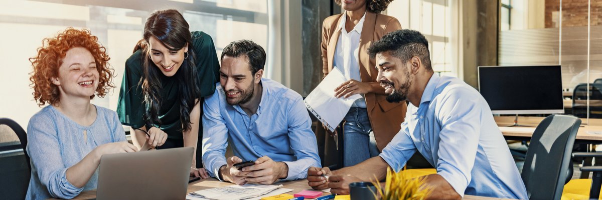 People gathered around one person in an office setting looking at something interesting on her laptop.