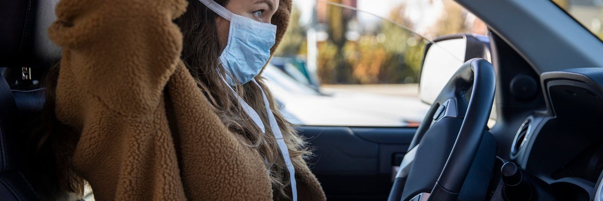 A person sits in the driver's seat of a parked vehicle and puts on a facemask.