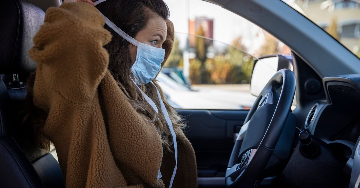 A person sits in the driver's seat of a parked vehicle and puts on a facemask.