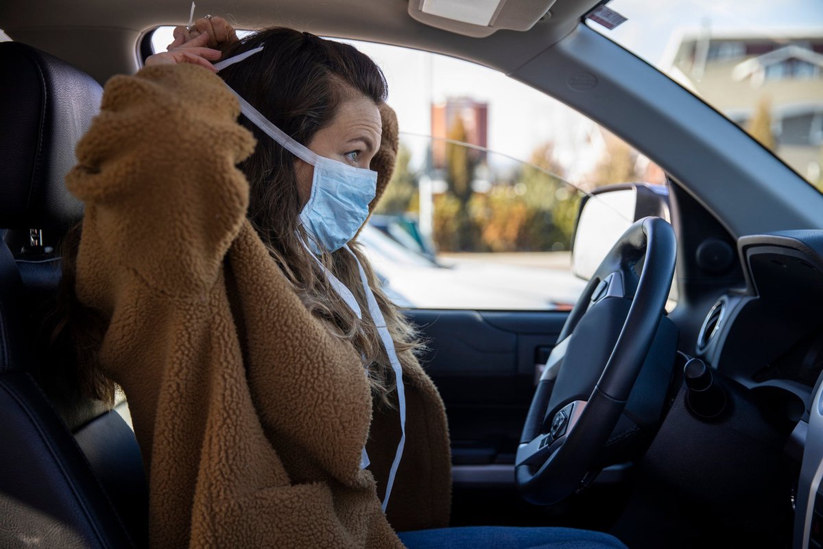 A person sits in the driver's seat of a parked vehicle and puts on a facemask.