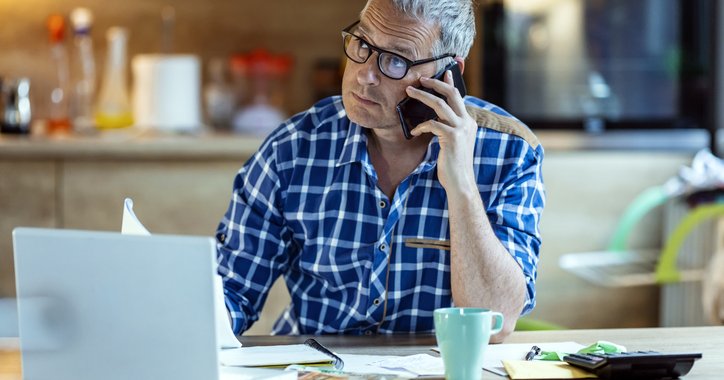 Worried man listens to a phone call while sitting in front of a laptop, calculator, and paperwork
