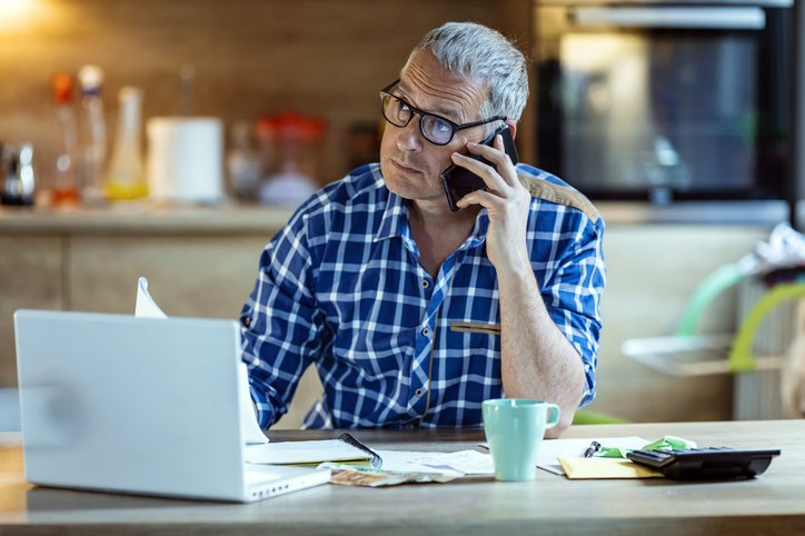 Worried man listening to a phone call while sitting in front of a laptop, calculator and paperwork