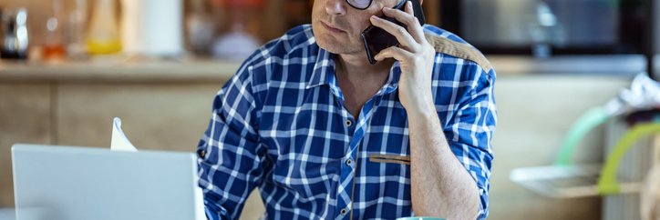 Worried man listens to a phone call while sitting in front of a laptop, calculator, and paperwork