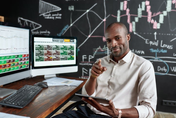 Man at desk with stocks and shares information smiles and points at camera.