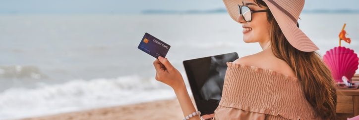 Woman wearing sun hat sits on a beach holding a credit card.