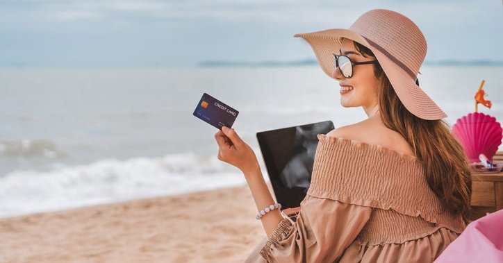 Woman wearing sun hat sits on a beach holding a credit card.