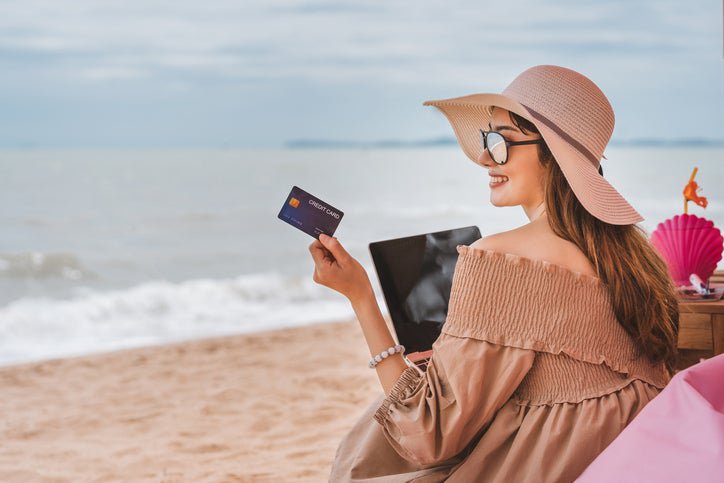 Woman wearing sun hat sits on a beach holding a credit card.