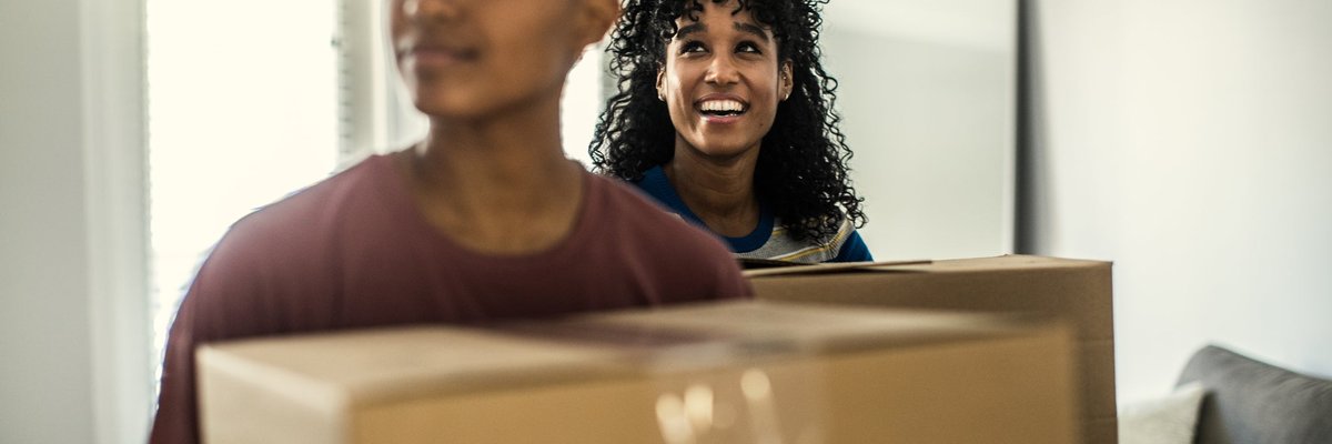 A couple smiles as they enter their new house carrying moving boxes.