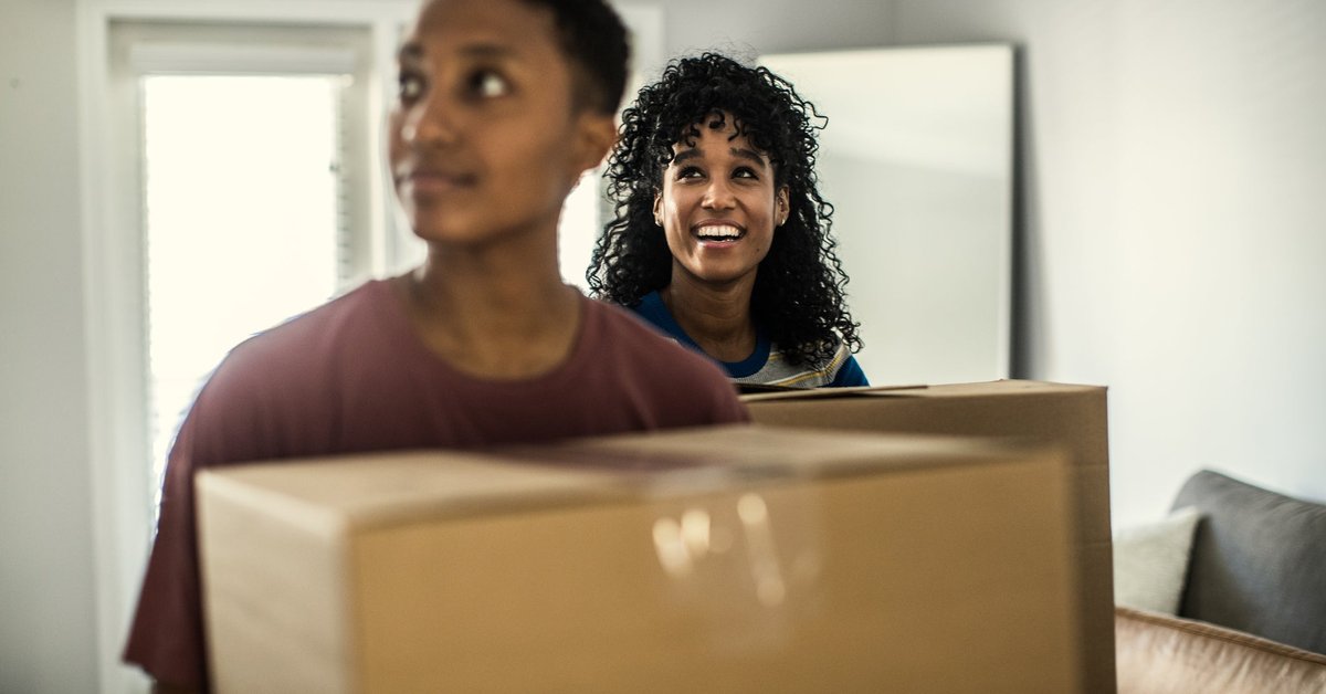 A couple smiles as they enter their new house carrying moving boxes.