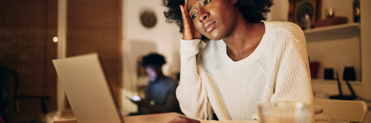 A person looks worriedly at their computer in their home, resting their hand on their head.