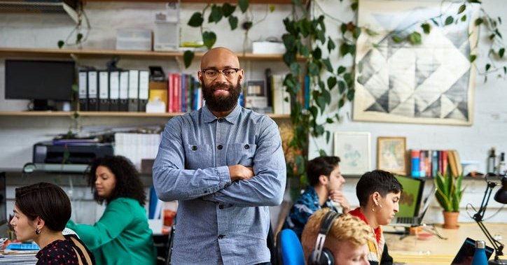 Bald person with beard stands in office with creative colleagues, smiling at camera.