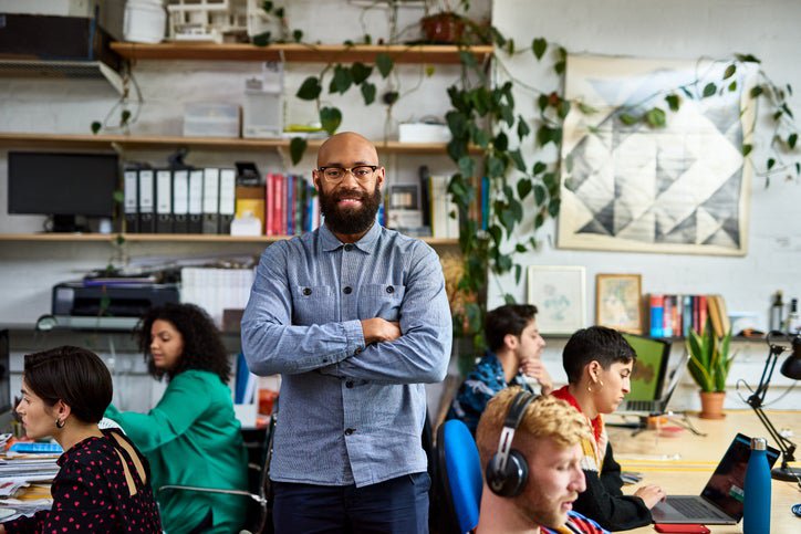 Bald person with beard stands in office with creative colleagues, smiling at camera.