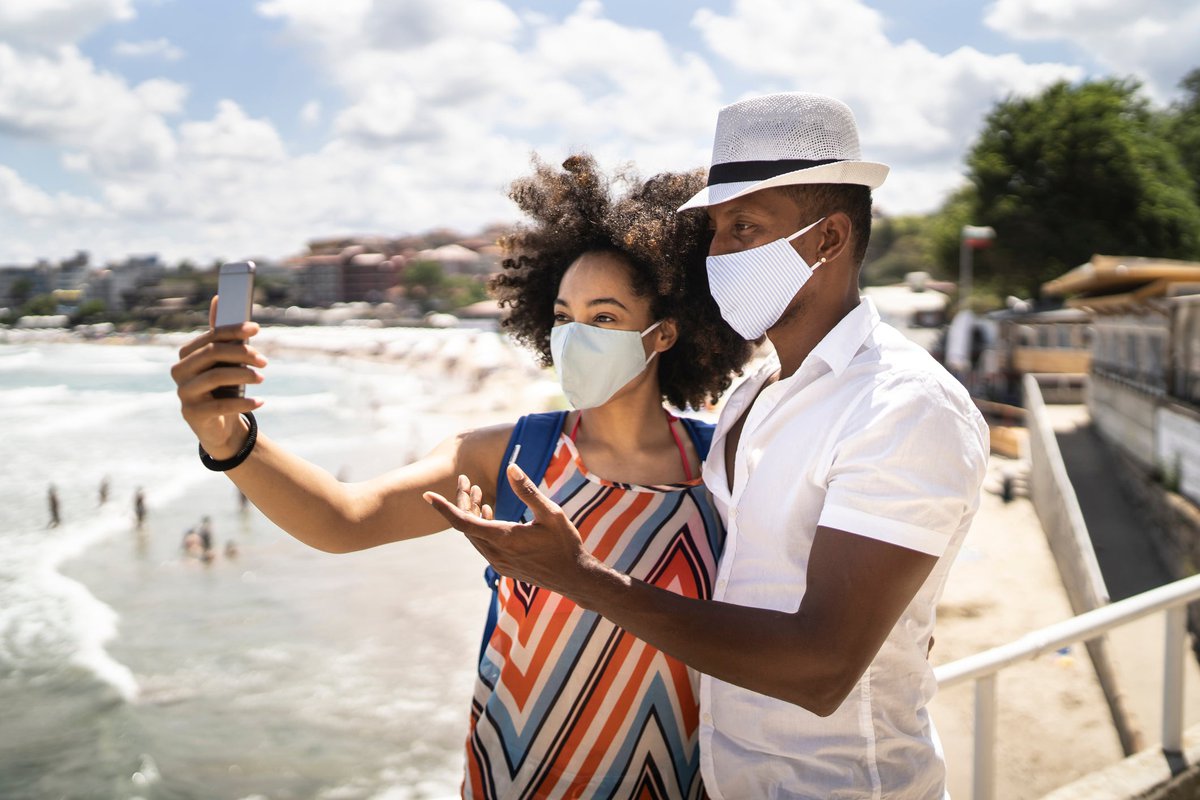 Young couple takes a selfie while wearing facemasks on the beach.