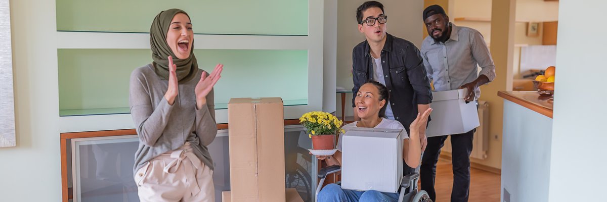 Four adults, including one using a wheelchair, gasp in delight at their new home surrounded by moving boxes.
