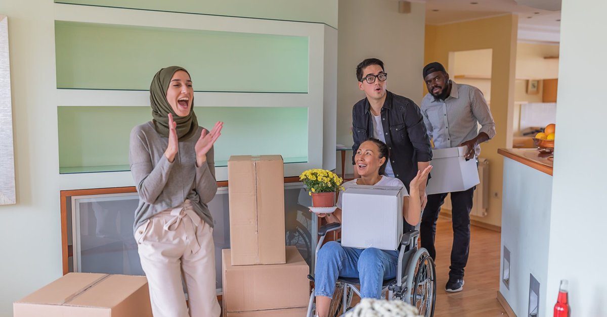 Four adults, including one using a wheelchair, gasp in delight at their new home surrounded by moving boxes.