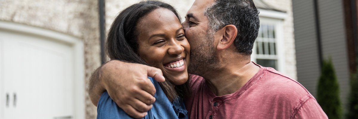 A couple happily embraces in front of a house, while one person kisses the other on the cheek.