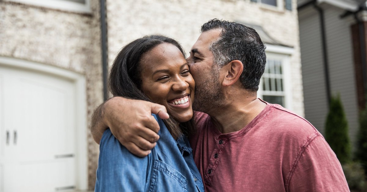 A couple happily embraces in front of a house, while one person kisses the other on the cheek.