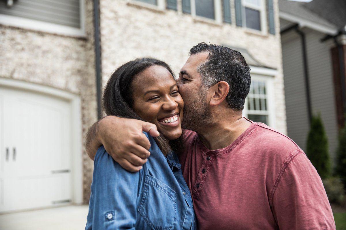 A couple happily embraces in front of a house, while one person kisses the other on the cheek.