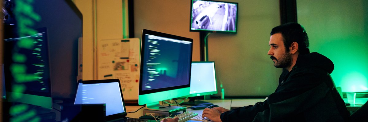 A hacker sits alone in a dark room, working on multiple computer screens.