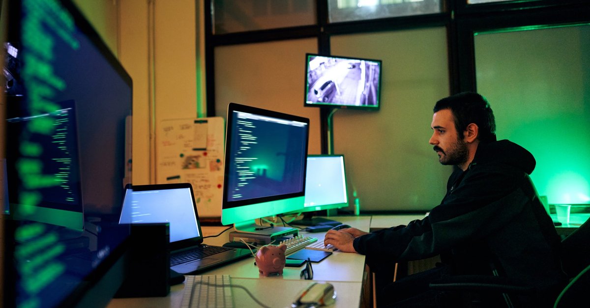A hacker sits alone in a dark room, working on multiple computer screens.