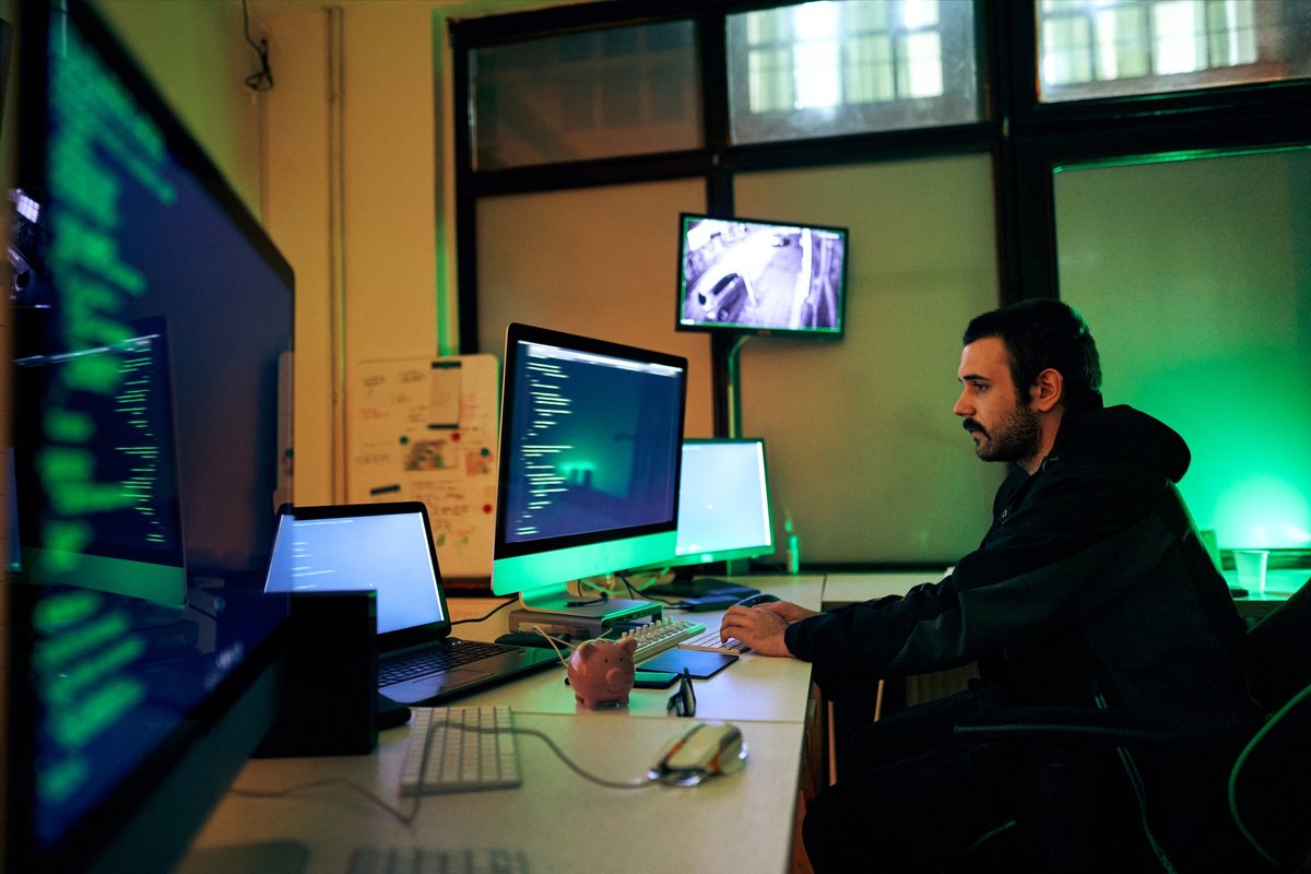 Un hacker est assis seul dans une pièce sombre, travaillant sur plusieurs écrans d'ordinateur.