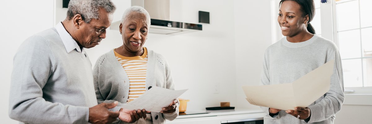 An insurance broker and an older couple discuss finances while standing in a kitchen holding folders.