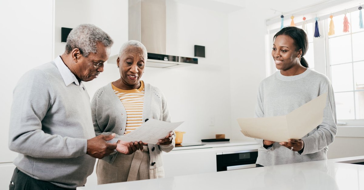 An insurance broker and an older couple discuss finances while standing in a kitchen holding folders.