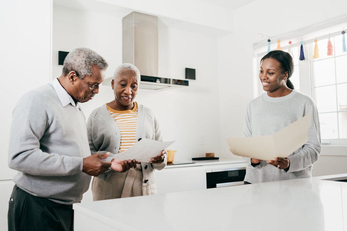 An insurance broker and an older couple discuss finances while standing in a kitchen holding folders.