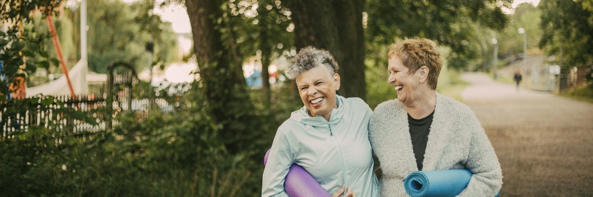 An older couple laugh with their arms around each other while holding yoga mats outside.