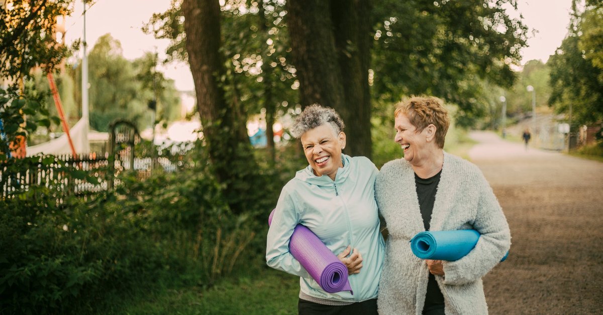 An older couple laugh with their arms around each other while holding yoga mats outside.